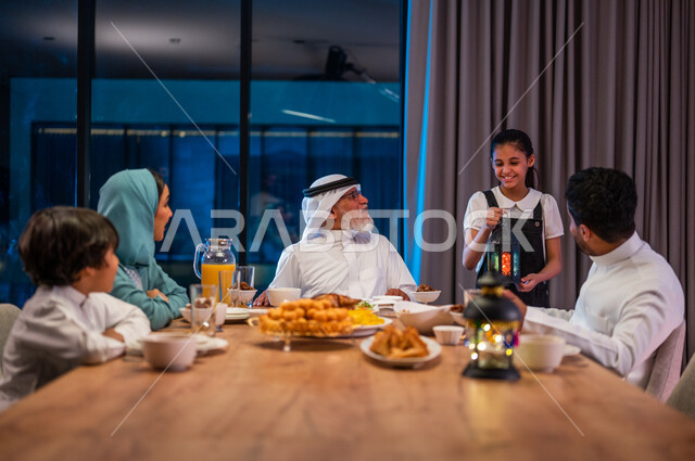 ديكورات زينة رمضانية، تبادل الأحاديث بين أفراد الأسرة، صورة مقربة لطفلة عربية خليجية سعودية تحمل بيدها فانوس معدني، اجتماع العائلة حول مائدة الطعام، سفرة مليئة باصناف من المأكولات الشرقية الشهية، أداء فريضة الصيام