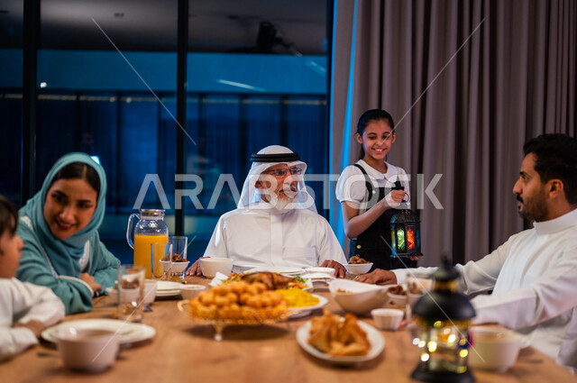 Exchanging conversations between family members, a religious Ramadan atmosphere, a close-up photo of a Saudi Gulf Arab girl holding a metal lantern in her hand, a family meeting around the dining table, a dinner filled with delicious oriental foods, performing the obligatory fasting.