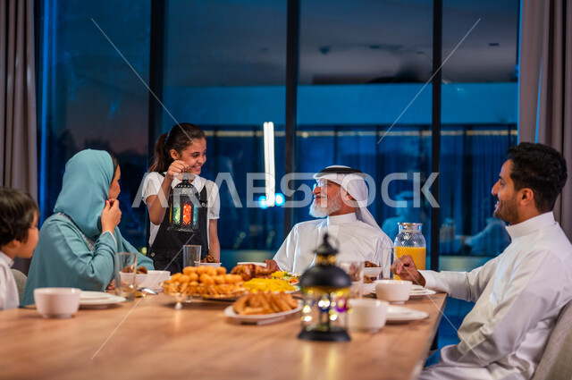 اجتماع العائلة حول مائدة الطعام، أداء فريضة الصيام، طفلة عربية خليجية سعودية تحمل بيدها فانوس معدني، ديكورات زينة رمضانية، تبادل الأحاديث بين أفراد الأسرة، سفرة مليئة باصناف من المأكولات الشرقية الشهية