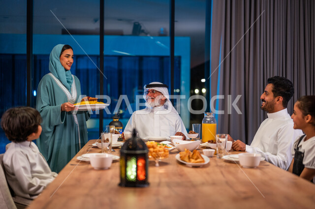 ايماءات الشكر والامتنان، امرأة محجبة ترتدي العباءة تقوم بإعداد مائدة الإفطار، اجتماع الأسرة العربية الخليجية السعودية حول طاولة الطعام المليئه بأصناف المأكولات و المقبلات الشهية، ديكورات وأجواء رمضانية