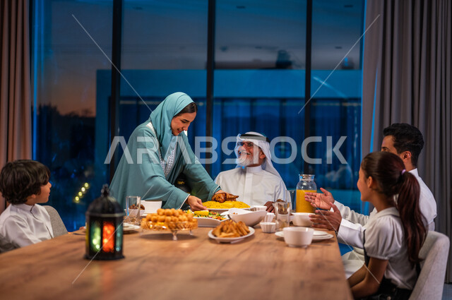 اجتماع الأسرة العربية الخليجية السعودية حول طاولة الطعام المليئه بأصناف المأكولات و المقبلات الشهية، ايماءات الشكر والامتنان، امرأة محجبة ترتدي العباءة تقوم بإعداد مائدة الإفطار، ديكورات وأجواء رمضانية