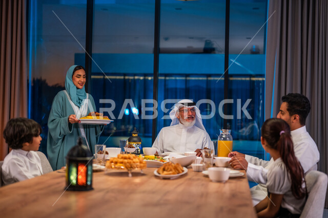 حوارات ونقاشات عائلية، امرأة محجبة ترتدي العباءة تقوم بتجهيز سفرة الإفطار، جتماع أسرة عربية خليجية سعودية حول مائده الطعام المليئه بأصناف المأكولات و المقبلات الشهية، ديكورات وأجواء رمضانية