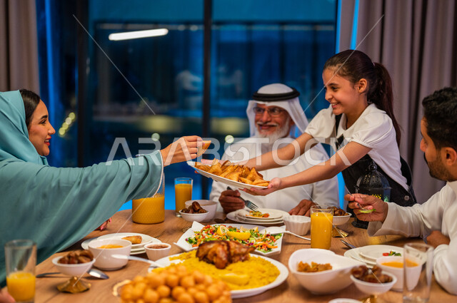 إكرام الوالدين ومشاركة الاطعمة، أجواء دينية رمضانية، صورة مقربة لطفلة عربية خليجية سعودية تقوم بتقريب الطبق لوالدتها، سفرة مليئة باصناف من المأكولات الشهية، اجتماع الأسرة حول مائدة الطعام، أداء فريضة الصيام