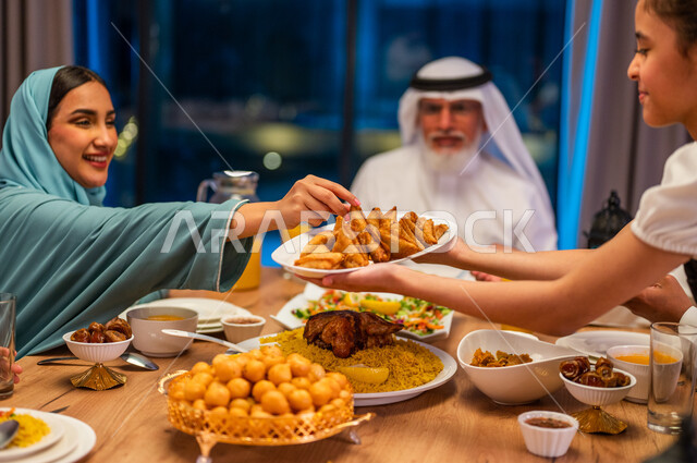 Gestures of happiness and joy, honoring one’s parents and sharing food, a religious Ramadan atmosphere, a close-up of a Saudi Gulf Arab girl bringing a plate to her mother, a dinner filled with delicious types of food, a family meeting around the dining table, performing the obligation of fasting.
