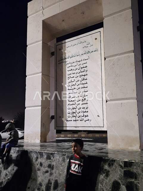 طفل عربي يقف أمام اللافتة التعريفية بأسماء شهداء غزوة بدر في المدينة المنورة في وضع الليل، الأحياء التراثية الشعبية الأثرية التاريخية، أماكن ومعالم إسلامية دينية مشهورة في المملكة العربية السعودية 