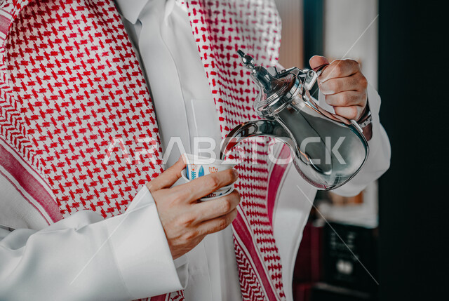 مفهوم كرم الضيافة وحسن الإستقبال، العادات والتقاليد الأصيلة, الاستمتاع بشرب البن العربي في الخيمة الشعبية، صورة مقربة ليد رجل عربي خليجي سعودي يرتدي الشماغ والثوب التقليدي يقوم بصب القهوة في الفنجان، التمسك بالقيم الكريمة المتوارثة من الأجداد
