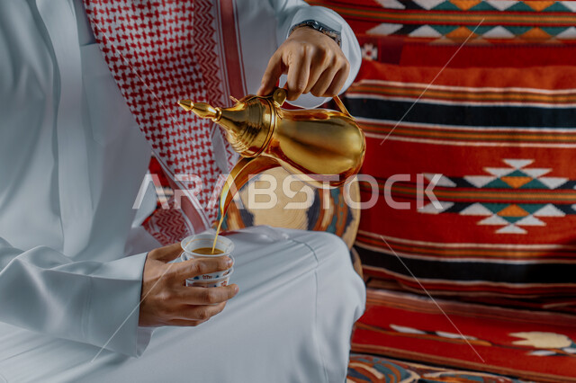مفهوم كرم الضيافة وحسن الإستقبال، العادات والتقاليد الأصيلة, الاستمتاع بشرب البن العربي في الخيمة الشعبية، صورة مقربة ليد رجل عربي خليجي سعودي يرتدي الشماغ والثوب التقليدي يقوم بصب القهوة في الفنجان، التمسك بالقيم الكريمة المتوارثة من الأجداد