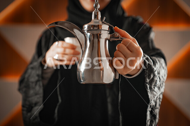 العادات والتقاليد الأصيلة, مفهوم كرم الضيافة وحسن الإستقبال، الاستمتاع بالمشروبات الساخنة، صورة مقربة لإمرأة عربية خليجية سعودية محجبة ترتدي العباءة السوداء تحمل بيدها دلة قهوة عربية وفناجين، التمسك بالقيم الكريمة المتوارثة من الأجداد