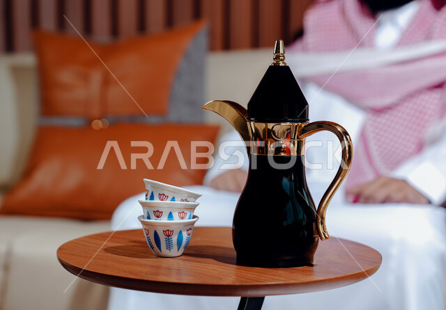 التمسك بالقيم الكريمة المتوارثة من الأجداد، مفهوم كرم الضيافة وحسن الإستقبال، العادات والتقاليد الأصيلة, صورة مقربة لدلة قهوة وفناجين على طاولة خشبية يجلس خلفها رجل عربي خليجي سعودي يرتدي الشماغ والثوب التقليدي يجلس على الكنبة، الاستمتاع بالمشروبات الساخنة