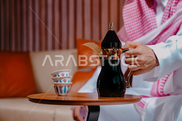 Adherence to noble values inherited from ancestors, the concept of hospitality and good reception, authentic customs and traditions, a close-up photo of a Saudi Gulf Arab man wearing a shemagh and traditional thobe, sitting on the sofa holding an Arabic coffee pot, enjoying hot drinks