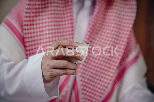  العادات والتقاليد السعودية الأصيلة, صورة مقربة لرجل عربي خليجي سعودي يرتدي الشماغ والثوب التقليدي يحمل بيده فنجان القهوة العربية، الاستمتاع بالمشروبات الساخنة، التمسك بالقيم الكريمة المتوارثة من الأجداد، مفهوم كرم الضيافة وحسن الإستقبال