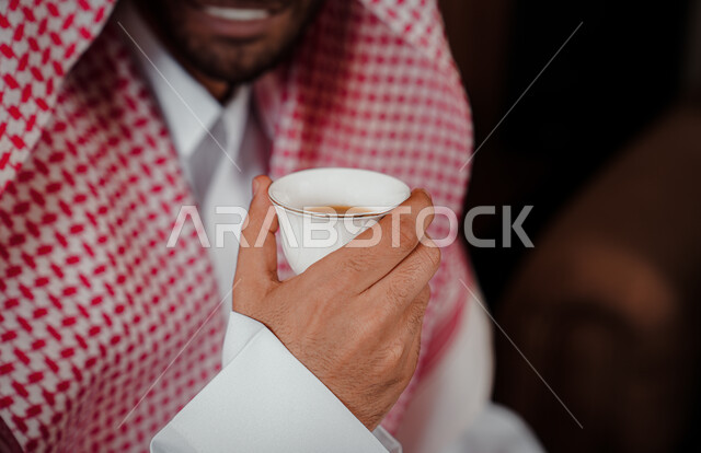 مفهوم كرم الضيافة وحسن الإستقبال، العادات والتقاليد السعودية الأصيلة, صورة مقربة لرجل عربي خليجي سعودي يرتدي الشماغ والثوب التقليدي يحمل بيده فنجان القهوة العربية، الاستمتاع بالمشروبات الساخنة، التمسك بالقيم الكريمة المتوارثة من الأجداد