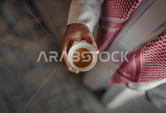 التمسك بالقيم الكريمة المتوارثة من الأجداد، مفهوم كرم الضيافة وحسن الإستقبال، العادات والتقاليد السعودية الأصيلة, صورة مقربة لرجل عربي خليجي سعودي يرتدي الشماغ والثوب التقليدي يحمل بيده فنجان القهوة العربية، الاستمتاع بالمشروبات الساخنة