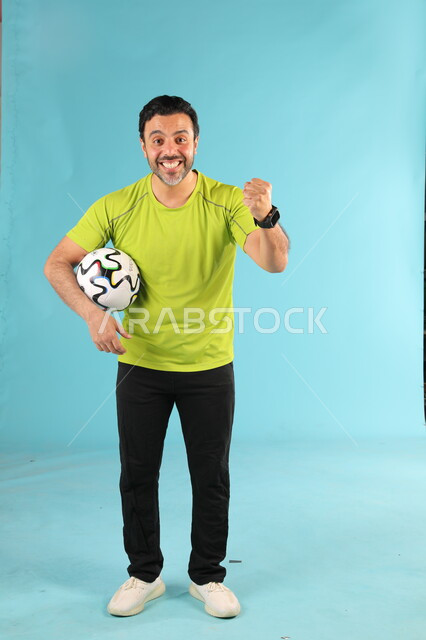 تعابير الفوز والانتصار، ممارسة الهوايات الرياضية، بورتريه لشاب عربي خليجي سعودي مبتسم يرتدي الملابس الرياضية يمسك بيده كرة القدم ينظر الى الكاميرا بايماءات السرور والحماس، الرياضات السعودية الشبابية، صورة الجسم بالطول الكامل، خلفية زرقاء
