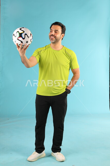 ممارسة الهوايات الرياضية، بورتريه لشاب عربي خليجي سعودي مبتسم يرتدي الملابس الرياضية يمسك بيده كرة القدم ينظر اليها بتعابير السرور والسعادة، الرياضات السعودية الشبابية، صورة الجسم بالطول الكامل، خلفية زرقاء