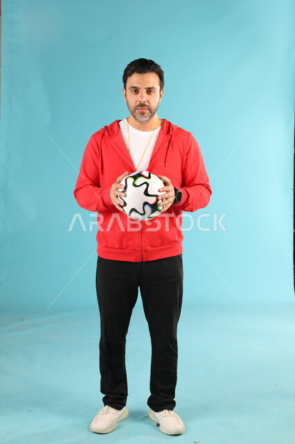 النظر الى الكاميرا بتعابير وايماءات الجدية والثقة بالنفس، الرياضات السعودية الشبابية، ممارسة الهوايات الرياضية، بورتريه لمدرب عربي خليجي سعودي يرتدي الملابس الرياضية يحمل كرة القدم يقف باستقامة  صورة الجسم بالطول الكامل، خلفية زرقاء