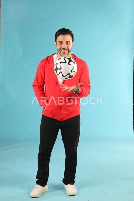 الوقوف باستقامة بايماءات وتعابير السرور والسعادة، الرياضات السعودية الشبابية، ممارسة الهوايات الرياضية، بورتريه لمدرب عربي خليجي سعودي مبتسم يرتدي الملابس الرياضية يقوم باللعب بكرة القدم، صورة الجسم بالطول الكامل، خلفية زرقاء
