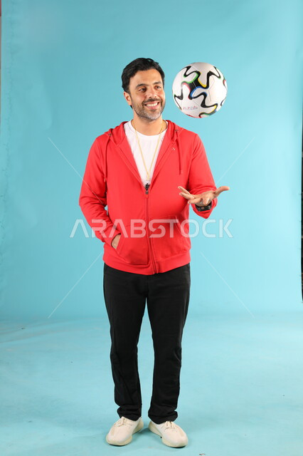 Standing upright with gestures and expressions of pleasure and happiness, Saudi youth sports, practicing sports hobbies, portrait of a smiling Saudi Arabian Gulf coach wearing sports clothes playing football, full-length body portrait, blue background