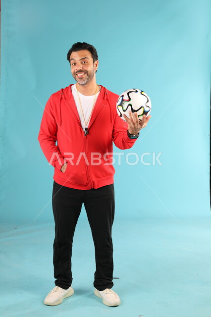  ممارسة الهوايات الرياضية، بورتريه لمدرب عربي خليجي سعودي مبتسم يرتدي الملابس الرياضية يمسك بيده كرة القدم يقف باستقامة ينظر الى الكاميرا بايماءات وتعابير السرور والسعادة، الرياضات السعودية الشبابية، صورة الجسم بالطول الكامل، خلفية زرقاء