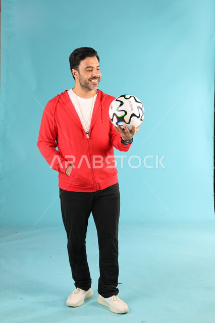 الوقوف باستقامة بايماءات وتعابير السرور والسعادة، الرياضات السعودية الشبابية، ممارسة الهوايات الرياضية، بورتريه لمدرب عربي خليجي سعودي مبتسم يرتدي الملابس الرياضية يقوم باللعب بكرة القدم، صورة الجسم بالطول الكامل، خلفية زرقاء