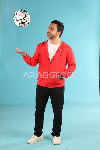 الوقوف باستقامة بايماءات وتعابير السرور والسعادة، الرياضات السعودية الشبابية، ممارسة الهوايات الرياضية، بورتريه لمدرب عربي خليجي سعودي مبتسم يرتدي الملابس الرياضية يقوم باللعب بكرة القدم، صورة الجسم بالطول الكامل، خلفية زرقاء
