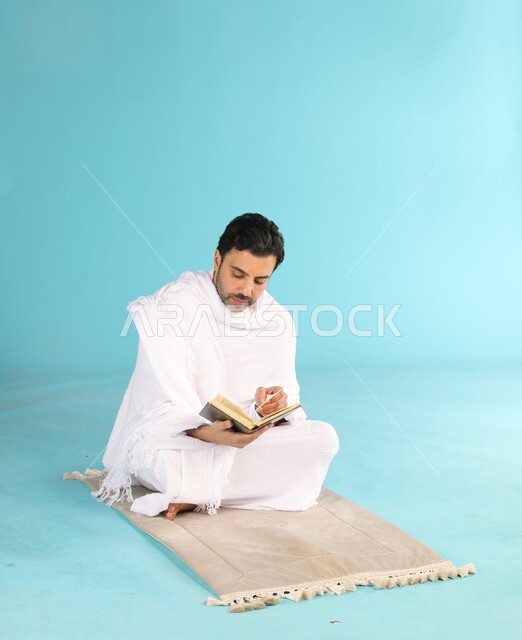 تلاوة وقراءة القرآن الكريم، بورتريه لرجل عربي سعودي خليجي يرتدي لباس الإحرام يحمل بيده المصحف الشريف يجلس على سجادة الصلاة, الشعور بالخشوع والطمأنينه, الإكثار من ذكر الله تعالى والإجتهاد في العباده, التقرب الى الله في شهر رمضان, خلفية زرقاء