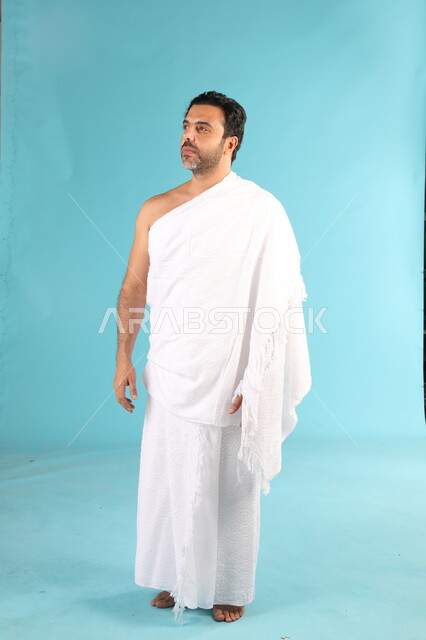 الاستعداد لأداء مناسك العمرة في شهر رمضان المبارك، بورتريه لرجل عربي خليجي سعودي مبتسم يرتدي لباس الاحرام يقوم بزيارة بيت الله الحرام في مكة المكرمة، العبادة والتقرب الى الله،، الوقوف بإستقامة والنظر الى شيء ما، صورة الجسم بالطول الكامل، خلفية زرقاء