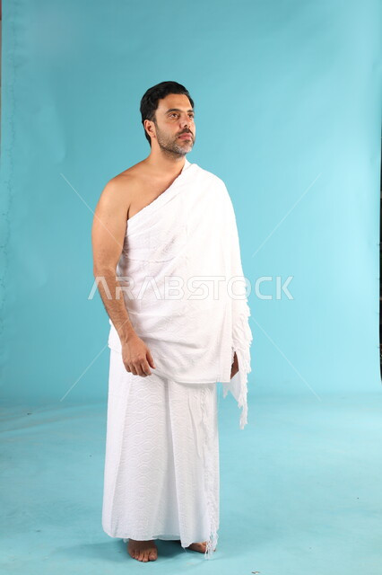 الوقوف بإستقامة والنظر الى شيء ما، الاستعداد لأداء مناسك العمرة في شهر رمضان المبارك، بورتريه لرجل عربي خليجي سعودي مبتسم يرتدي لباس الاحرام يقوم بزيارة بيت الله الحرام في مكة المكرمة، العبادة والتقرب الى الله، صورة الجسم بالطول الكامل، خلفية زرقاء