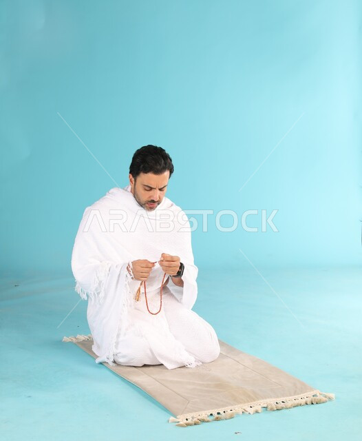 مفهوم التسبيح والتهليل، الإجتهاد في العباده والتقرب الى الله في شهر رمضان المبارك, بورتريه لرجل عربي سعودي خليجي يرتدي لباس الاحرام يجلس على سجادة الصلاة يمسك بيده السبحه, الإكثار من ذكر الله تعالى من خلال الإستغفار والدعاء, خلفيه زرقاء