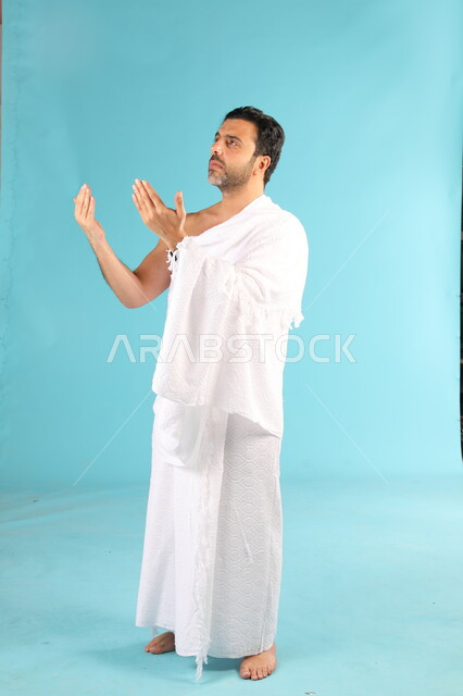الإكثار من الدعاء في شهر رمضان المبارك، التقرب الى الله من خلال الإجتهاد في العبادة والحفاظ الطاعات، بورتريه لرجل عربي سعودي خليجي يرتدي لباس الإحرام يرفع يديه ويدعو الله بإيماءات تدل على التوسل والتضرع، صورة الجسم بالكامل، خلفية زرقاء