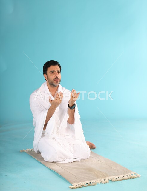 أداء فروض الصلاة على وقتها, التقرب الى الله من خلال الإجتهاد في العبادة واداء مناسك الحج والعمرة، بورتريه لرجل عربي سعودي خليجي يرتدي لباس الاحرام يجلس على سجادة الصلاة يرفع يديه ويدعو الله، إيماءات تدل على التوسل والتضرع، خلفية زرقاء