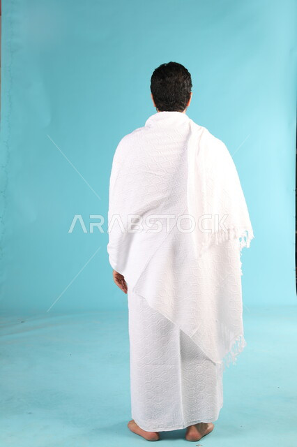 الطاعة والعبادة والتقرب الى الله، الوقوف بإستقامة والنظر الى الكاميرا، الاستعداد لأداء مناسك العمرة في شهر رمضان المبارك، بورتريه من الخلف لرجل عربي خليجي سعودي يرتدي لباس الاحرام يقوم بزيارة بيت الله الحرام في مكة المكرمة، صورة الجسم بالطول الكامل، خلفية زرقاء