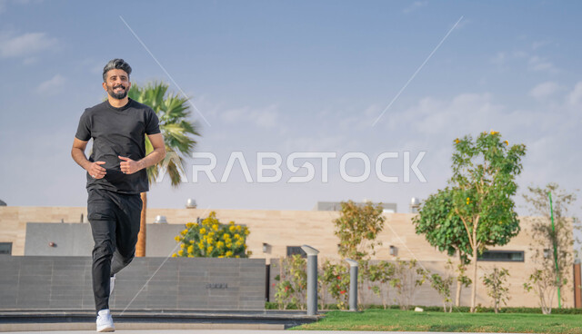 النباتات الخضراء والأشجار، رجل عربي خليجي سعودي مبتسم يقوم بالتمارين الصباحية، الصحه واللياقه الجسديه، نشاط بدني في الهواء الطلق، ايماءات السعادة والسرور، التنزه في الحدائق العامة، ممارسة رياضة المشي في ممشى المنتزه، صورة لكامل الجسم