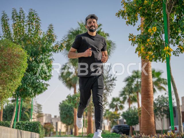 منظر الطبيعة في الحدائق و المنتزهات العامة، ممارسة رياضة المشي، النباتات الخضراء والأشجار، رجل عربي خليجي سعودي مبتسم يقوم بالتمارين الصباحية، الصحه واللياقه الجسديه، نشاط بدني في الهواء الطلق، ايماءات السعادة والسرور، صورة لكامل الجسم