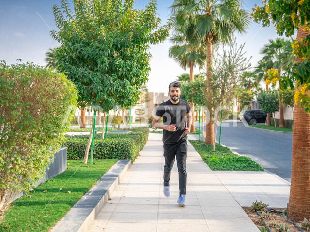 التنزه في الحدائق العامة، نشاط بدني في الهواء الطلق، ممارسة رياضة المشي في ممشى المنتزه، الصحه واللياقه الجسديه، رجل عربي خليجي سعودي يقوم بالتمارين الصباحية، الأشجار والنباتات الخضراء، صورة لكامل الجسم