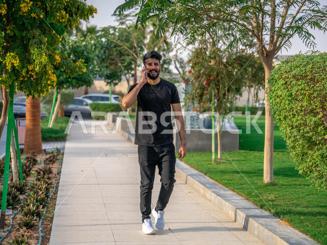 اجراء المكالمات الهاتفية، النباتات الخضراء والأشجار، التنزه في الحدائق العامة، نشاط بدني في الهواء الطلق، ممارسة رياضة المشي في ممشى المنتزه، الصحه واللياقه الجسديه، رجل عربي خليجي سعودي يقوم بالتمارين الصباحية، صورة لكامل الجسم