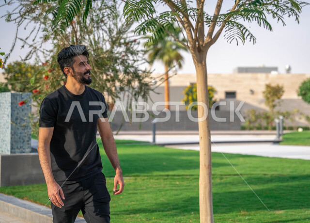 ايماءات السعادة والسرور، نشاط بدني في الهواء الطلق، الصحه واللياقه الجسديه، النباتات الخضراء والأشجار، التنزه في الحدائق العامة، ممارسة رياضة المشي في ممشى المنتزه، رجل عربي خليجي سعودي مبتسم يقوم بالتمارين الصباحية