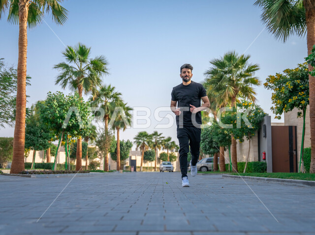 التنزه في الحدائق العامة، نشاط بدني في الهواء الطلق، ممارسة رياضة المشي في ممشى المنتزه، الصحه واللياقه الجسديه، رجل عربي خليجي سعودي يقوم بالتمارين الصباحية، الأشجار والنباتات الخضراء، صورة لكامل الجسم