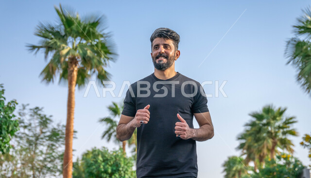 تعابير السعادة والسرور، النباتات الخضراء والأشجار، التنزه في الحدائق العامة، نشاط بدني في الهواء الطلق، ممارسة رياضة المشي في ممشى المنتزه، الصحه واللياقه الجسديه، رجل عربي خليجي سعودي مبتسم يقوم بالتمارين الصباحية