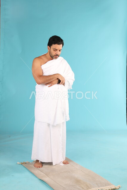 Performing the obligatory prayers on time, getting closer to God through diligence in worship and performing the rituals of Hajj and Umrah, a portrait of a Saudi Gulf Arab man wearing the Ihram dress praying on the prayer rug, gestures indicating pleading and pleading, full-length body image, blue background.