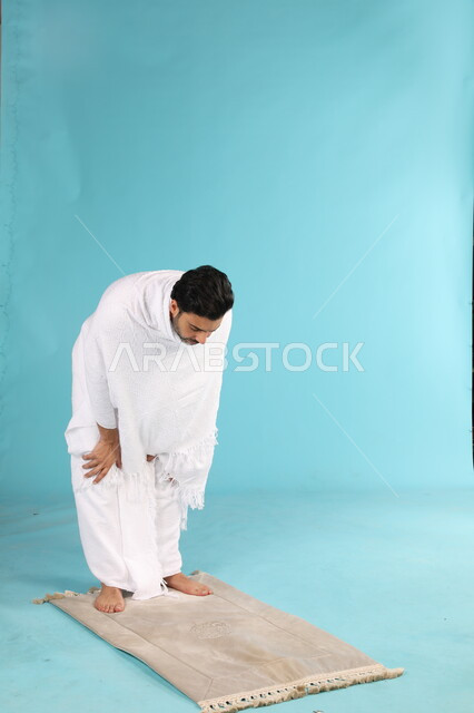 أداء فروض الصلاة على وقتها, التقرب الى الله من خلال الإجتهاد في العبادة واداء مناسك الحج والعمرة، بورتريه لرجل عربي سعودي خليجي يرتدي لباس الاحرام يركع على سجادة الصلاة، إيماءات تدل على التوسل والتضرع، صورة الجسم بالطول الكامل، خلفية زرقاء