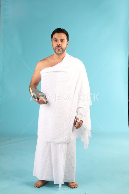 قراءة وتلاوة القرآن الكريم، بورتريه لرجل عربي سعودي خليجي يرتدي لباس الإحرام يحمل بيده المصحف الشريف، الشعور بالخشوع والطمأنينه, الإكثار من ذكر الله تعالى والإجتهاد في العباده, التقرب الى الله في شهر رمضان المبارك, صورة الجسم بالكامل، خلفية زرقاء