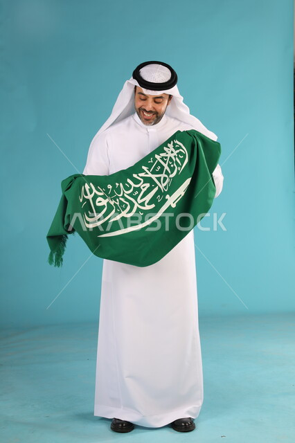  احياء ذكرى اليوم الوطني السعودي 23 سبتمبر، بورتريه لرجل عربي خليجي سعودي مبتسم يرتدي الغترة والثوب التقليدي  يحمل شعار السعودية للاحتفال في يوم العلم 11 مارس، مناسبات وطنية شعبيه، اظهار الحب والولاء للمملكه، صورة الجسم بالطول الكامل، خلفية زرقاء
