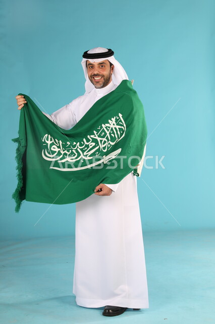  احياء ذكرى اليوم الوطني السعودي 23 سبتمبر، بورتريه لرجل عربي خليجي سعودي مبتسم يرتدي الغترة والثوب التقليدي  يحمل شعار السعودية للاحتفال في يوم العلم 11 مارس، مناسبات وطنية شعبيه، اظهار الحب والولاء للمملكه، صورة الجسم بالطول الكامل، خلفية زرقاء