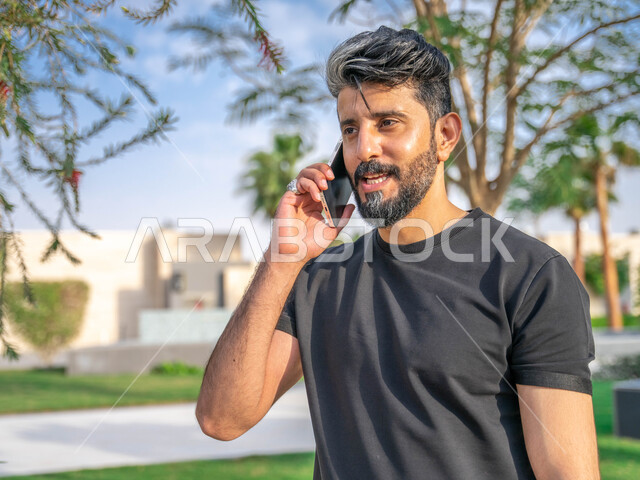 تبادل الاحاديث المضحكة والمسلية عبرالموبايل، المنتزهات والحدائق العامة، اجراء مكالمات الهاتفية اثناء ممارسة رياضة المشي، الدردشة والتواصل مع الأهل والاصدقاء، صورة مقربة لرجل عربي خليجي سعودي مبتسم يقوم بالتمارين الصباحية، نشاط بدني في الهواء الطلق