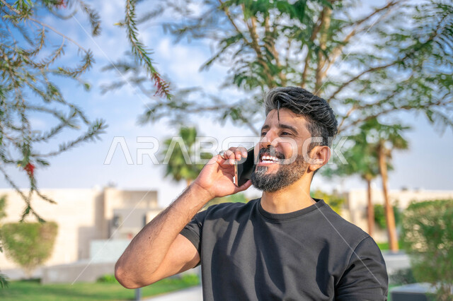 المنتزهات والحدائق العامة، الدردشة والتواصل مع الأهل والاصدقاء، تبادل الاحاديث المضحكة والمسلية عبرالموبايل، الصحه واللياقه الجسديه، اجراء مكالمات الهاتفية اثناء ممارسة رياضة المشي، صورة مقربة من الجانب لرجل عربي خليجي سعودي مبتسم يقوم بالتمارين الصباحية