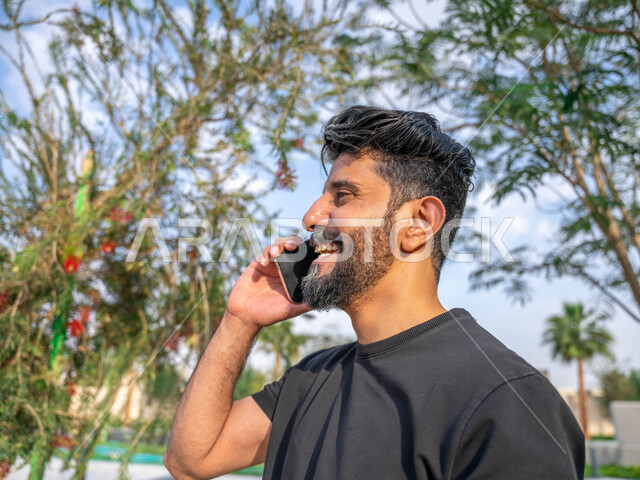 الدردشة والتواصل مع الأهل والاصدقاء، تبادل الاحاديث المضحكة والمسلية عبرالموبايل، الصحه واللياقه الجسديه، اجراء مكالمات الهاتفية اثناء ممارسة رياضة المشي، المنتزهات والحدائق العامة، صورة مقربة من الجانب لرجل عربي خليجي سعودي مبتسم يقوم بالتمارين الصباحية