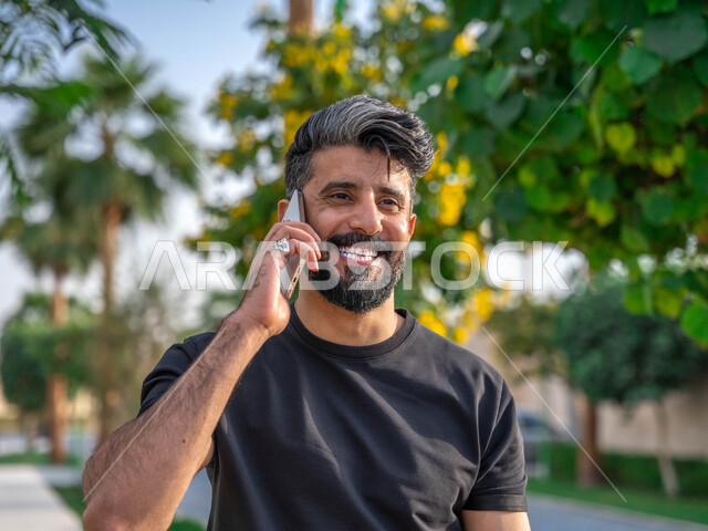 اجراء مكالمات الهاتفية اثناء ممارسة رياضة المشي، تبادل الاحاديث المضحكة والمسلية عبرالموبايل، المنتزهات والحدائق العامة، الدردشة والتواصل مع الأهل والاصدقاء، صورة مقربة لرجل عربي خليجي سعودي مبتسم يقوم بالتمارين الصباحية