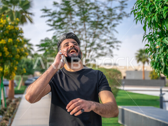 تبادل الاحاديث المضحكة والمسلية عبرالموبايل، المنتزهات والحدائق العامة، اجراء مكالمات الهاتفية اثناء ممارسة رياضة المشي، الدردشة والتواصل مع الأهل والاصدقاء، صورة مقربة لرجل عربي خليجي سعودي مبتسم يقوم بالتمارين الصباحية، نشاط بدني في الهواء الطلق