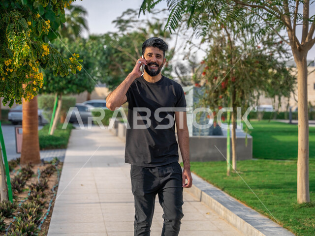 اجراء مكالمات الهاتفية اثناء ممارسة رياضة المشي، تبادل الاحاديث المضحكة والمسلية عبرالموبايل، الدردشة والتواصل مع الأهل والاصدقاء، المنتزهات والحدائق العامة، رجل عربي خليجي سعودي مبتسم يقوم بالتمارين الصباحية، الصحه واللياقه الجسديه، نشاط بدني في الهواء الطلق